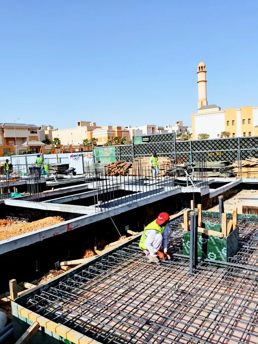 عامل بخوذة حمراء على شبكة تسليح قاعدة خرسانية، وفي الخلفية مئذنة.